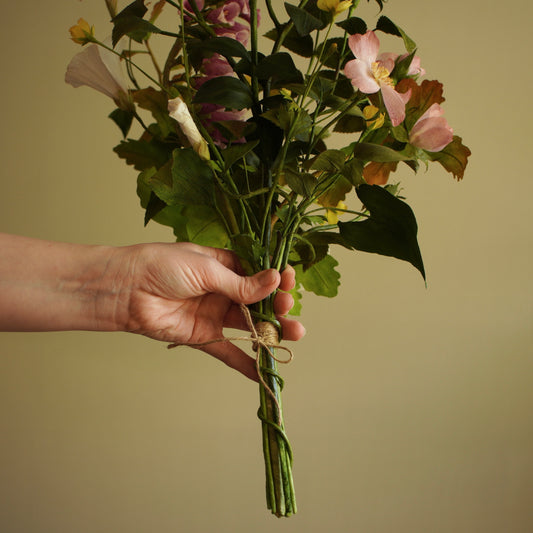A hand holding a bouquet of artificial flowers, including pink dog rose, white bindweed and green foliage, tied with twine against a plain beige background.