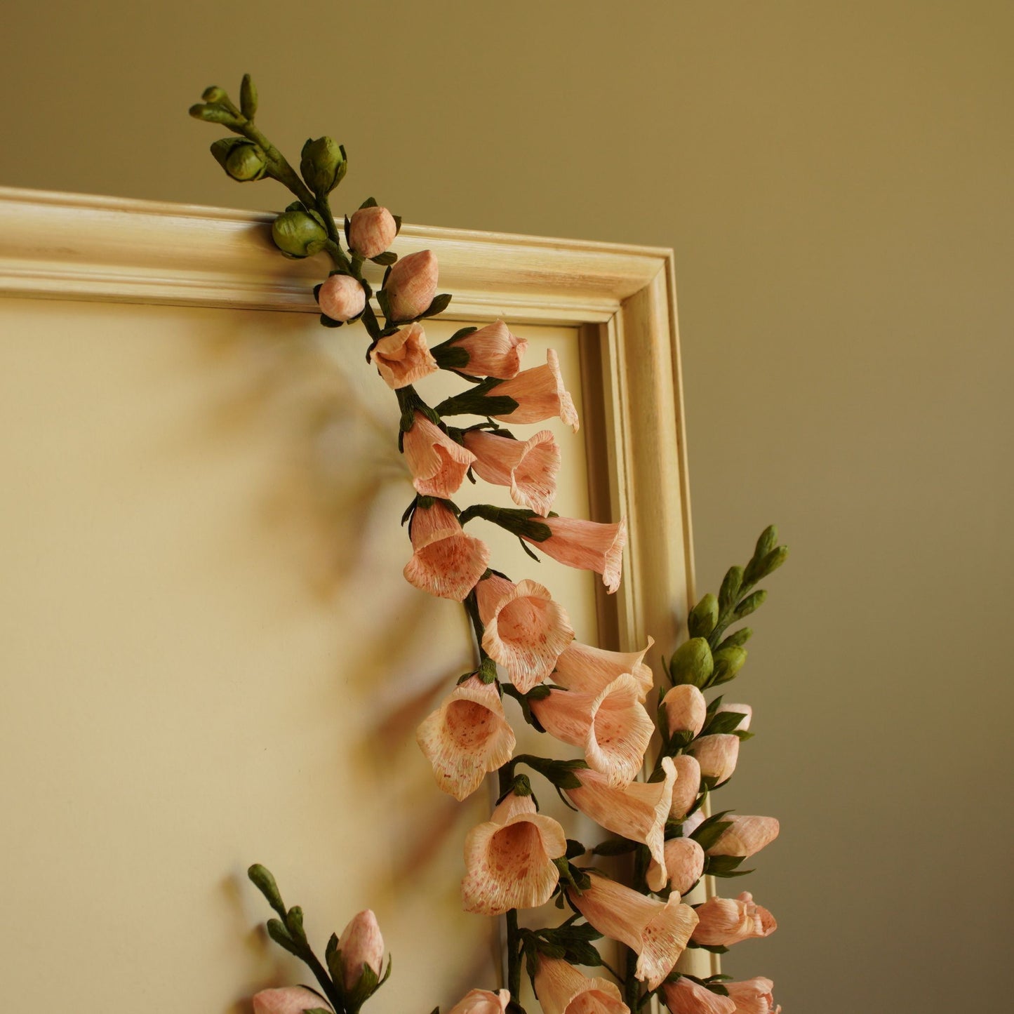 Upper section of framed paper foxglove artwork with gently arching stem and unopened buds, showing corner of painted and aged frame