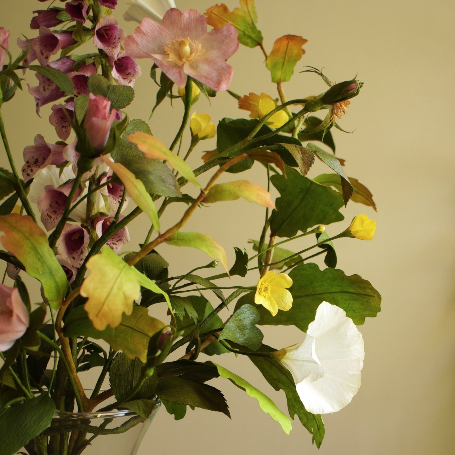 A mixed artificial flower arrangement featuring purple foxglove,  yellow buttercups, white bindweed and green foliage on a beige background.