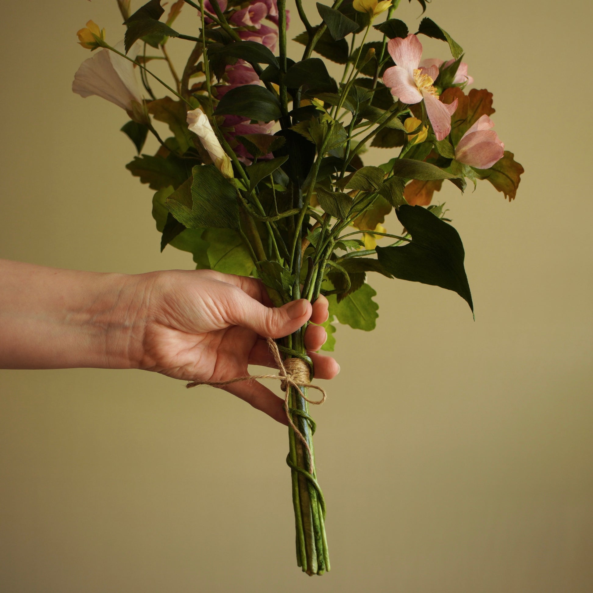 A hand holding a bouquet of artificial flowers, including pink dog rose, white bindweed and green foliage, tied with twine against a plain beige background.