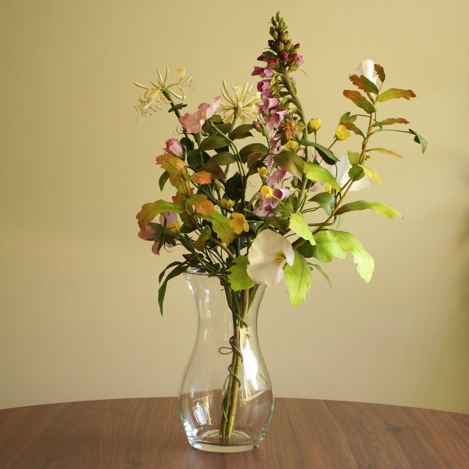 A mixed floral arrangement in a clear glass vase, featuring white bindweed, pink and purple flowers, and green foliage, set on a wooden table against a plain beige wall.