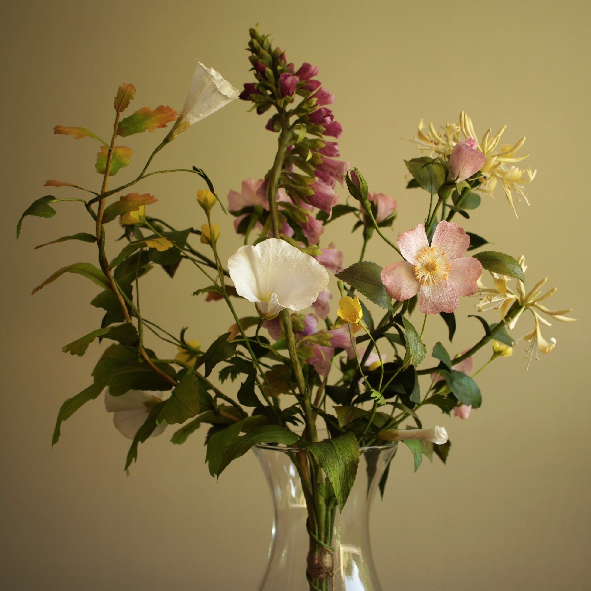 A mixed floral arrangement made from paper in a clear glass vase against a plain beige background. The bouquet includes white and pale pink flowers, purple blossoms, green leaves, and yellow accents.