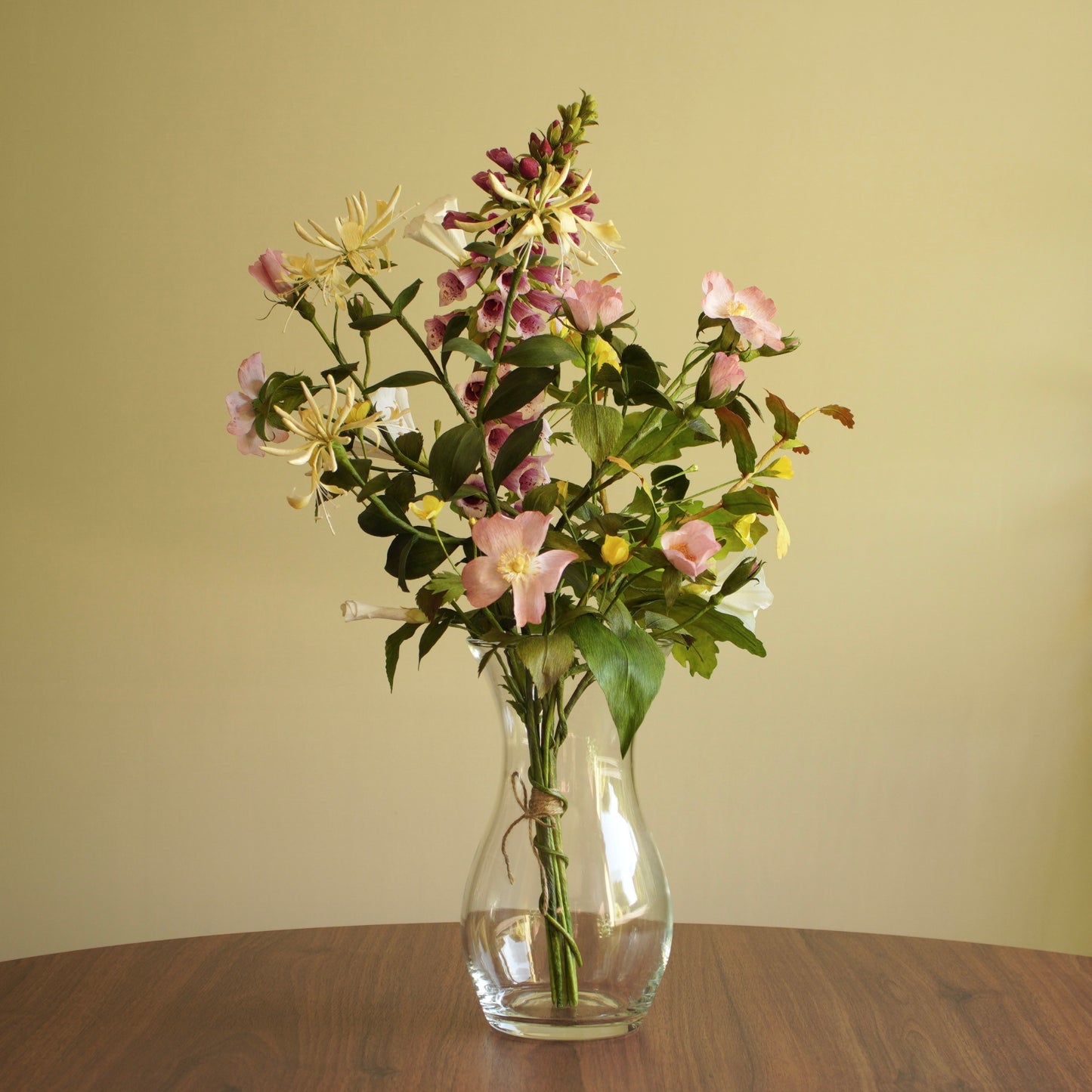 A mixed artificial flower bouquet with pink, white, and yellow blossoms and green leaves, arranged in a clear glass vase on a wooden table against a beige wall.