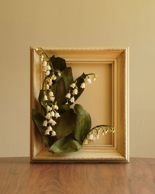 Full view of framed paper lily of the valley botanical artwork, arranged within a painted frame on a wooden surface in warm natural light