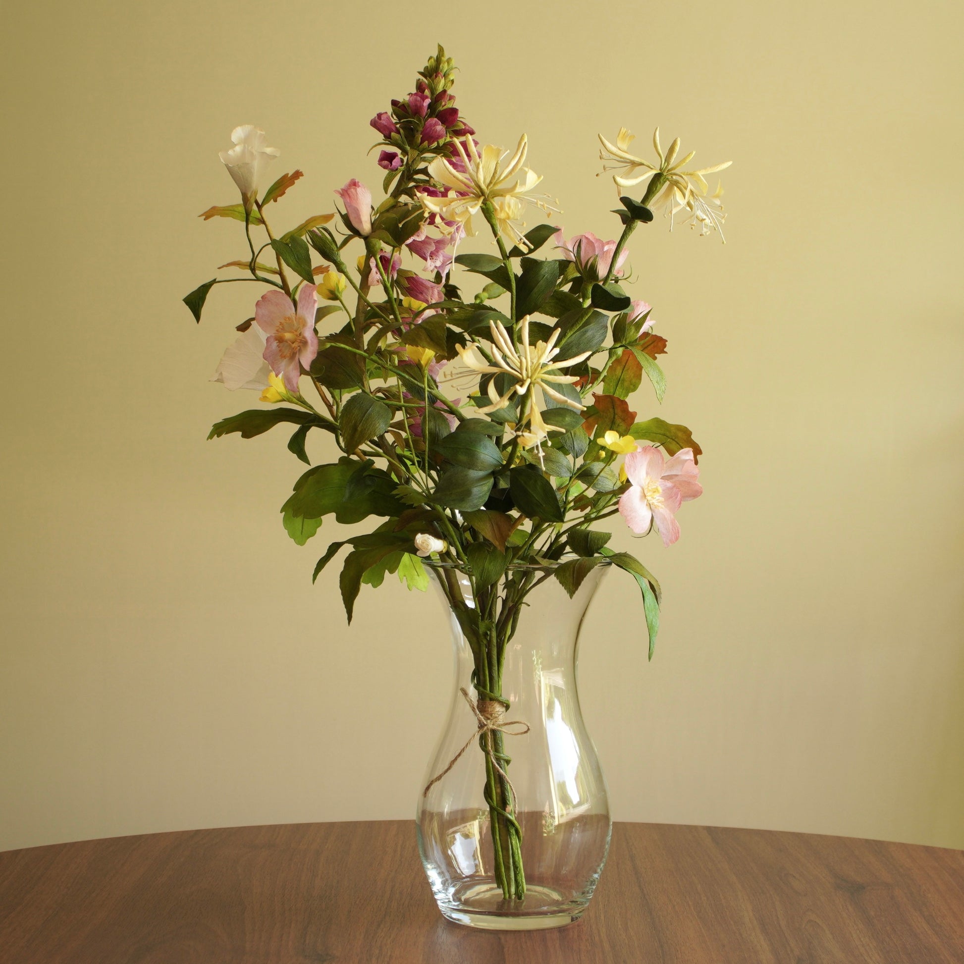 A mixed artificial flower bouquet made from paper, with green foliage, pink blossoms, and cream flowers arranged in a clear glass vase on a wooden table against a beige wall.