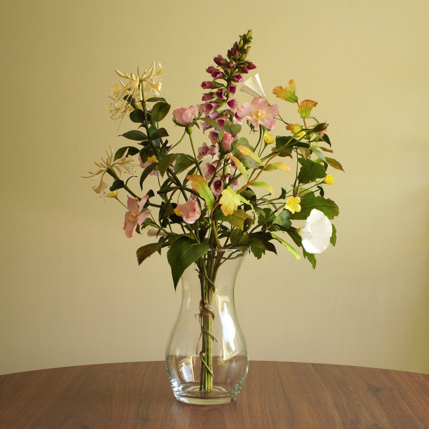 A mixed artificial flower arrangement in a clear glass vase, placed on a wooden table against a plain beige wall.