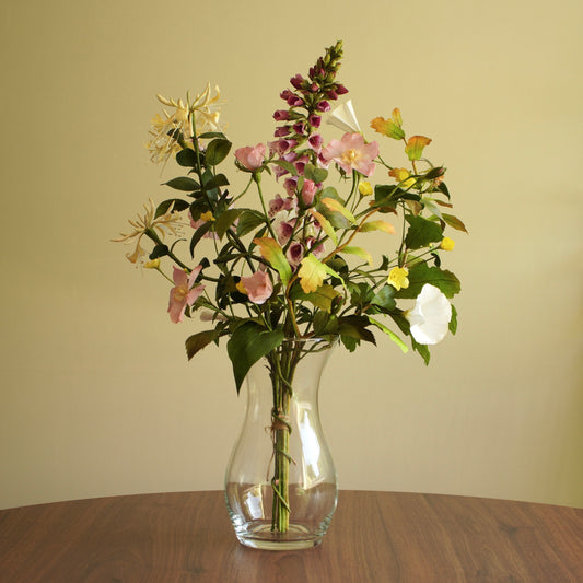 A mixed artificial flower arrangement in a clear glass vase, placed on a wooden table against a plain beige wall.