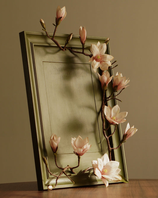 Framed paper magnolia branch with sculptural blooms arranged across painted green frame on wooden surface