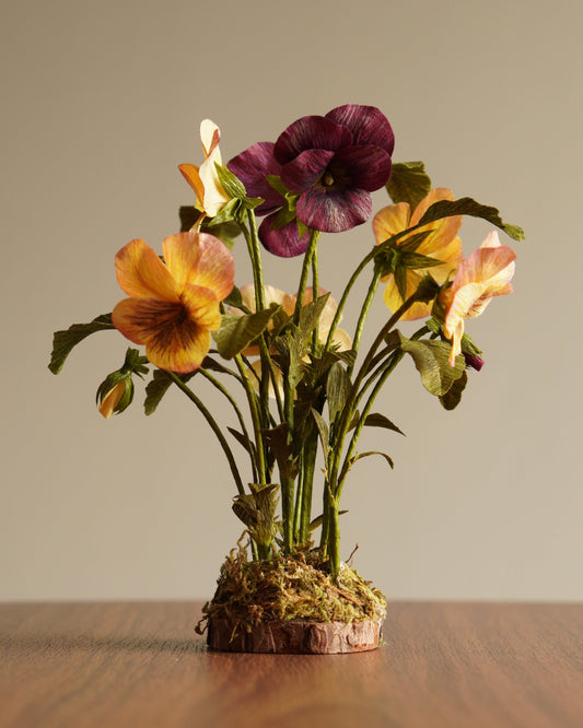 Paper pansy botanical sculpture in yellow and purple on moss-covered wood base on wooden surface